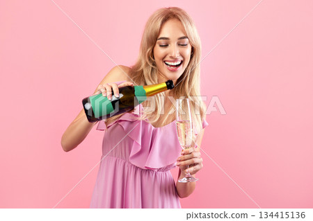 Cheerful blonde lady pouring champagne into glass over pink studio background, free space. Gorgeous young woman celebrating birthday, Valentine's Day, having hen party. Festive concept 134415136