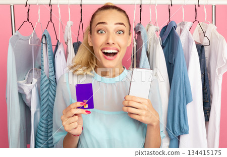 Cheerful Buyer. Excited overjoyed woman holding smartphone and purple credit card in hands, ordering stylish clothes and purchases online via internet, standing with rack in the pink background 134415175