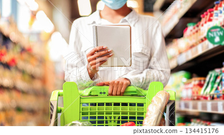 Unrecognizable Female Buyer Holding Grocery Shopping Checklist Buying Food Products, Standing With Full Shop Cart In Supermarket Groceries Store. Woman Consumer Choosing Healthy Food Concept. Cropped 134415176
