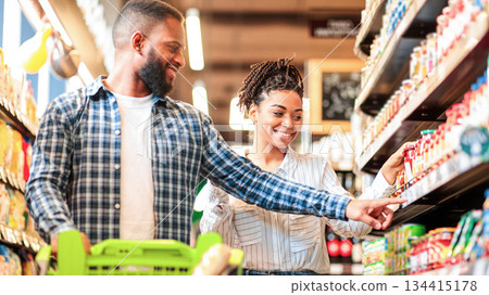 African American Spouses Buying Food In Grocery Shop Pointing Fingers At Products Walking With Cart Full Of Goods Standing In Supermarket Indoors. Family Buying Groceries Together Concept African American Spouses Buying Food In Grocery Shop Pointing Fingers At Products Walking With Cart Full Of Goods Standing In Supermarket Indoors. Family Buying Groceries Together Concept 134415178