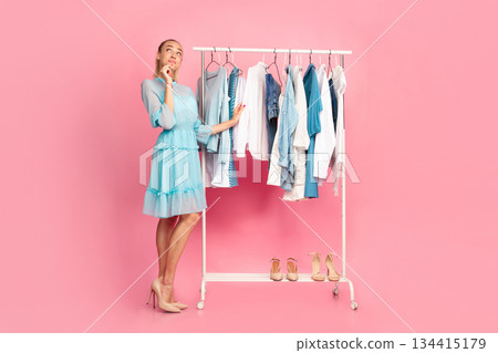 Making Choice. Vertical full length portrait of young pensive lady choosing trendy outfit, thinking about purchase and what to wear, standing in wardrobe near clothing rail, pink background Making Choice. Vertical full length portrait of young pensive lady choosing trendy outfit, thinking about purchase and what to wear, standing in wardrobe near clothing rail, pink background 134415179