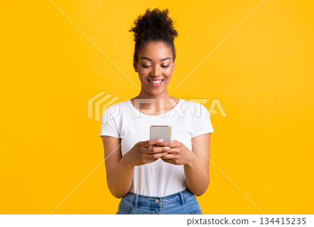 People, Technology And Communication Concept. Portrait of beautiful black lady texting on her cell phone, isolated over yellow studio background, copy space. Smiling woman chatting in social media People, Technology And Communication Concept. Portrait of beautiful black lady texting on her cell phone, isolated over yellow studio background, copy space. Smiling woman chatting in social media 134415235