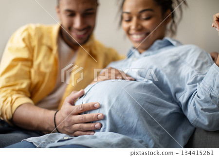 Pregnancy. Joyful Black Spouses Expecting Baby, Husband Touching Wife's Belly Sitting On Couch At Home. Family And Childbirth Concept. Cropped, Selective Focus 134415261