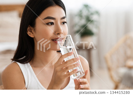 Hydration concept. Young attractive asian woman drinking spring water at home, looking at copy space. Beautiful lady holding glass of mineral water, hydrating her body in the morning 134415280