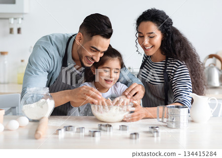 Cute arab family baking pastry together at kicthen, copy space. Joyful middle-eastern father, mother and little daughter making dough for home-made cookies. Domestic family lifestyle concept Cute arab family baking pastry together at kicthen, copy space. Joyful middle-eastern father, mother and little daughter making dough for home-made cookies. Domestic family lifestyle concept 134415284