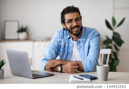Freelance Career. Handsome Indian Guy In Casual Wear Smiling At Camera While Sitting At Laptop Desk In Home Office, Young Cheerful Western Man Enjoying Working Remotely Online On Computer, Copy Space 134415291