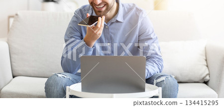 Smiling Businessman Talking By Phone In Loudspeaker Mode Or Using Voice Assistant On Smartphone Sitting At Laptop Computer On Couch Indoors. Gadgets And Modern Lifestyle, Voice Search Application Smiling Businessman Talking By Phone In Loudspeaker Mode Or Using Voice Assistant On Smartphone Sitting At Laptop Computer On Couch Indoors. Gadgets And Modern Lifestyle, Voice Search Application 134415295