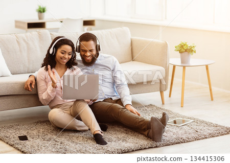 Online Video Call. Smiling balck couple having remote chat with friends or family using laptop, wearing headphones, waving hand to webcam, sitting on the floor carpet in living room at home 134415306
