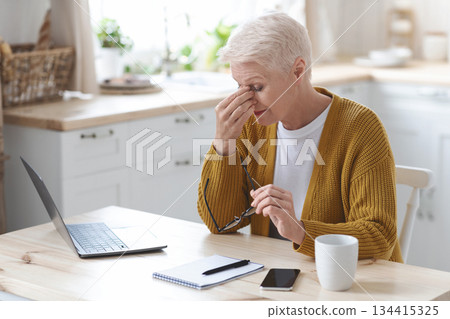Tired mature grey-haired woman freelancer working from home, using laptop, sitting in kitchen, copy space. Senior lady taking notes, sitting in front of computer, holding glasses, rubbing nose bridge 134415325