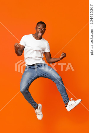 Cute black guy winner jumping up in the air and showing funny figures, having good day, showing positive emotions over orange background. African american man posing at studio, having fun Cute black guy winner jumping up in the air and showing funny figures, having good day, showing positive emotions over orange background. African american man posing at studio, having fun 134415367
