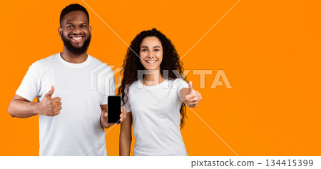 Smiling Multiethnic Couple Showing Cellphone Empty Screen Approving Application Gesturing Thumbs Up Standing Over Yellow Studio Background. We Like This Mobile App. Mockup, Panorama With Copy Space Smiling Multiethnic Couple Showing Cellphone Empty Screen Approving Application Gesturing Thumbs Up Standing Over Yellow Studio Background. We Like This Mobile App. Mockup, Panorama With Copy Space 134415399