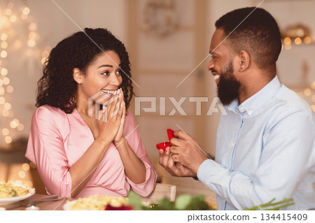 Be My Wife. Portrait of happy black man holding and giving open box with engagement ring to his surprised excited girlfriend, asking to marry him in cafe or at home during romantic date 134415409