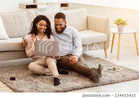 Online Communication. Smiling african american man and woman making digital video chat with friends or family using laptop, waving to webcam, sitting on the floor carpet in living room 134415433