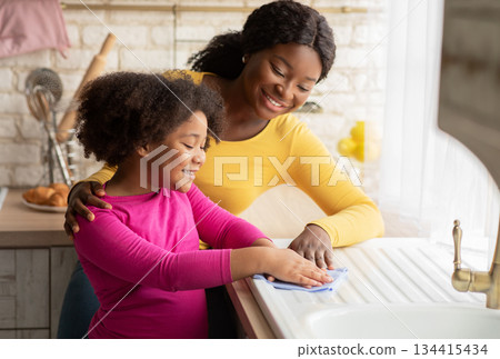 Loving Black Mother Teaching Her Daughter Housekeeping, Cleaning Kitchen Together. Cute Little Girl Wiping Table With Cloth And Smiling, Enjoying Helping With Household Chores, Free Space 134415434