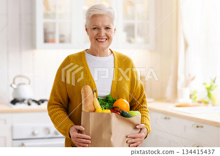 Happy mature woman holding a paper bag full of groceries from the supermarket, kitchen interior. Elderly woman with groceries at home, food delivery during quarantine concept, copy space 134415437