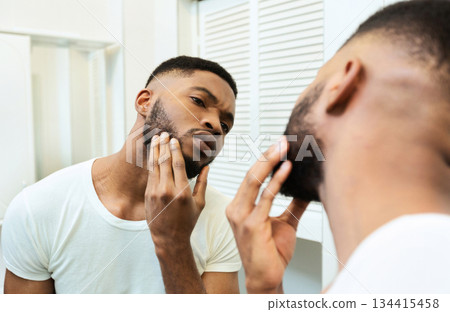Need to shave. Concentrated african guy looking at his beard at bathroom mirror Need to shave. Concentrated african guy looking at his beard at bathroom mirror 134415458