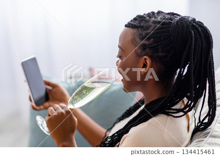 Pretty black woman with dreadlocks drinking champagne while using smartphone with blank screen, mockup, copy space. African american lady celebrating St. Valentines Day with her lover online 134415461