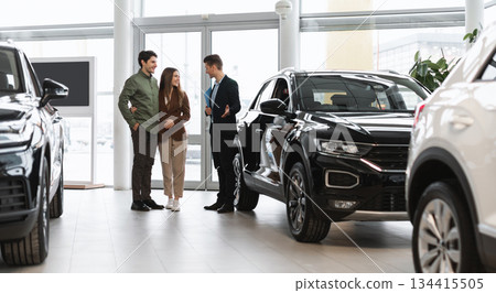 Car purchase or rental. Positive young spouses speaking to salesman about buying new auto at dealership, panorama. Successful millennial manager showing clients choice of vehicles at salon 134415505