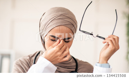Closeup of female doctor woman in hijab removing glasses and rubbing nose bridge, suffering from tired eyes syndrome, clinic interior. Eye fatigue, symptoms and treatments concept Closeup of female doctor woman in hijab removing glasses and rubbing nose bridge, suffering from tired eyes syndrome, clinic interior. Eye fatigue, symptoms and treatments concept 134415508