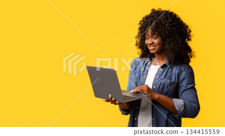 Pretty young black lady using modern laptop on yellow studio background, copy space. Cute african american curly woman chatting with friends or checking webstores. Modern technologies concept Pretty young black lady using modern laptop on yellow studio background, copy space. Cute african american curly woman chatting with friends or checking webstores. Modern technologies concept 134415559