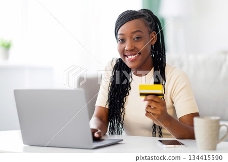 Smiling african american young woman holding credit card, using laptop at home, copy space. Happy black lady shopping online, ordering food, cosmetics or clothes from internet while staying home Smiling african american young woman holding credit card, using laptop at home, copy space. Happy black lady shopping online, ordering food, cosmetics or clothes from internet while staying home 134415590
