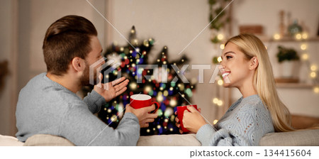 Christmas couple in love sitting on couch, drinking cocoa and having conversation in living room, copy space. Young married happy man and woman enjoying winter holidays together at home, back view 134415604