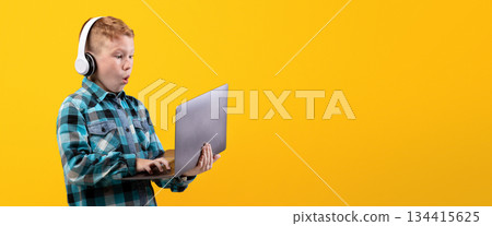Excited redhead boy with wireless headset playing on laptop, yellow studio background. Amazed kid in casual outfit using modern powerful gadget for video games, playing online, copy space Excited redhead boy with wireless headset playing on laptop, yellow studio background. Amazed kid in casual outfit using modern powerful gadget for video games, playing online, copy space 134415625