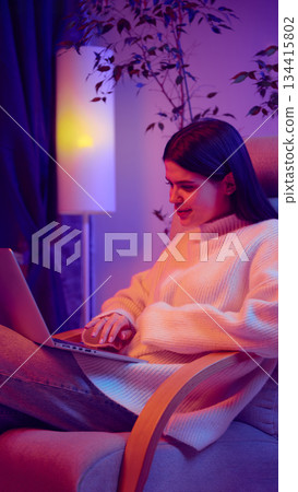Young woman working on laptop in armchair at home. 134415802
