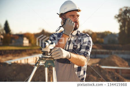 On the radio, with geodetic optical level. Worker is on the construction site at daytime 134415988
