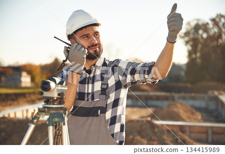 On the radio, with geodetic optical level. Worker is on the construction site at daytime 134415989