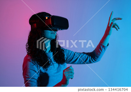 A woman immersed in virtual reality raises her hand toward digital space in neon light 134416704