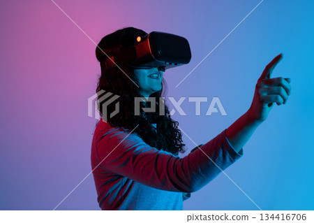 A woman wearing a virtual reality headset reaches forward and interacts with digital elements in soft neon blue and pink lighting. 134416706