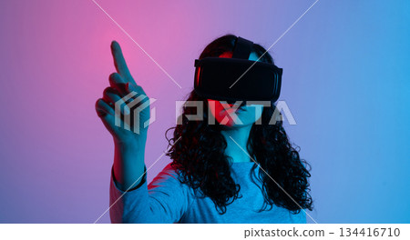 A woman in a VR headset raises her hand and points upward while exploring a digital environment under neon lighting. A woman in a VR headset raises her hand and points upward while exploring a digital environment under neon lighting. 134416710