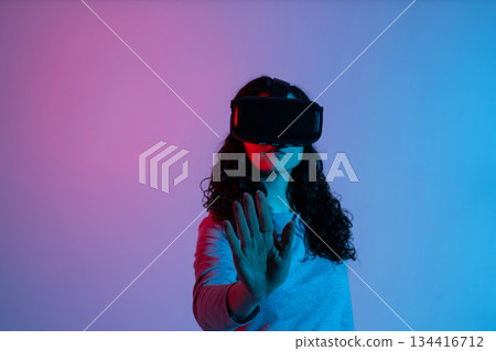 A woman wearing VR goggles extends her hand to interact with a virtual environment A woman wearing VR goggles extends her hand to interact with a virtual environment 134416712
