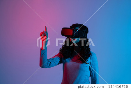 Woman using VR headset with neon blue red lighting while interacting with virtual interface in studio scene 134416726
