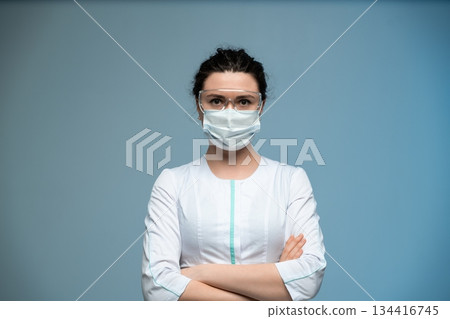 Female doctor in medical mask and glasses posing confidently with folded arms on blue background 134416745