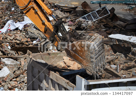 Excavator bucket loading debris into a dump truck, illustrating demolition, construction, and waste management process on a building site. Excavator bucket loading debris into a dump truck, illustrating demolition, construction, and waste management process on a building site. 134417097