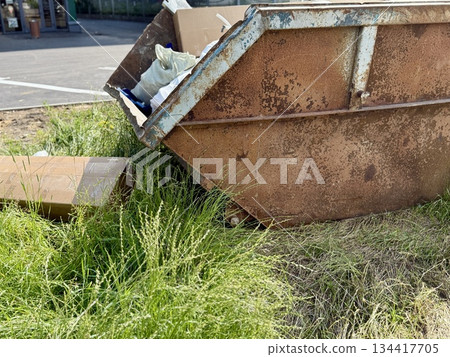 An Old and Rusty Dump Container That is Surrounded by Lush, Overgrown Grass and Weeds. A weathered metal dumpster overflowing with various debris, positioned against a backdrop of lush tall grass. An Old and Rusty Dump Container That is Surrounded by Lush, Overgrown Grass and Weeds. A weathered metal dumpster overflowing with various debris, positioned against a backdrop of lush tall grass. 134417705