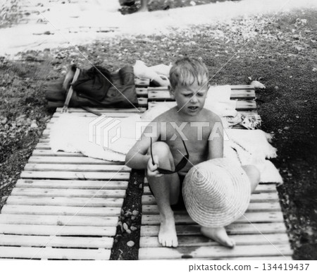 Vintage portrait of cute, blond boy wearing sunglasses and a straw hat on a wooden beach chair. Retro photo from 1971. Vintage portrait of cute, blond boy wearing sunglasses and a straw hat on a wooden beach chair. Retro photo from 1971. 134419437
