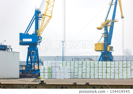 Industrial harbor with blue yellow cranes standing stacked packaged timber cargo port terminal. Maritime logistics scene highlights export operations heavy machinery structured waterfront industry 134419545