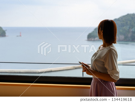 一位女士望著窗外的大海，手裡拿著她的智慧型手機。 134419618