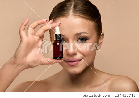 Smiling young white woman with bare shoulders standing over beige background, holding bottle of facial cosmetic oil. Cosmetic procedures for facial skin care. 134419715