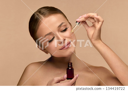 Woman Dropping Serum on Face on Beige Background. European Girl Applying Serum Collagen Moisturizer from Pipette on Face close up. Positive Laughing Beauty Woman Portrait. Concept of Face Skin Care Woman Dropping Serum on Face on Beige Background. European Girl Applying Serum Collagen Moisturizer from Pipette on Face close up. Positive Laughing Beauty Woman Portrait. Concept of Face Skin Care 134419722