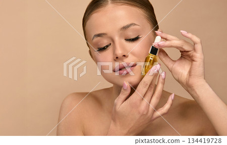 Smiling young white woman with bare shoulders standing over beige background, holding bottle of facial cosmetic oil. Cosmetic procedures for facial skin care Smiling young white woman with bare shoulders standing over beige background, holding bottle of facial cosmetic oil. Cosmetic procedures for facial skin care 134419728