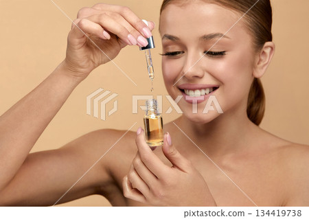 Beautiful young woman applying serum onto her face on beige background Beautiful young woman applying serum onto her face on beige background 134419738