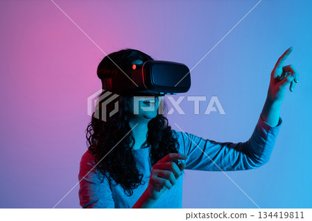 A woman raises her arm to interact with digital objects inside a VR space filled with neon blue and pink illumination. A woman raises her arm to interact with digital objects inside a VR space filled with neon blue and pink illumination. 134419811