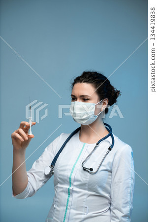 Nurse holding syringe inspecting dosage while wearing protective mask and uniform in clinical setting 134419838