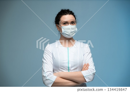 Female medical worker with protective mask and glasses posing with folded arms on blue background 134419847
