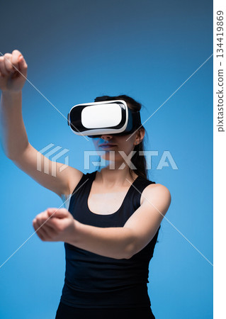 Young woman using a VR headset and moving her hands forward while interacting with a virtual environment in cool blue light Young woman using a VR headset and moving her hands forward while interacting with a virtual environment in cool blue light 134419869