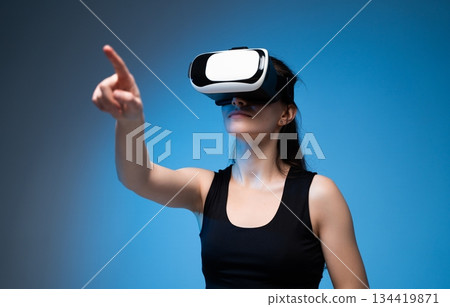 Woman in VR headset pointing forward while exploring immersive virtual reality world in blue studio Woman in VR headset pointing forward while exploring immersive virtual reality world in blue studio 134419871